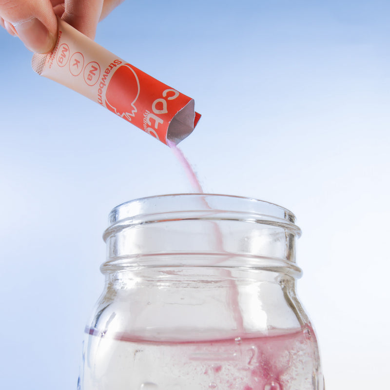Hand pouring electrolyte powder from a strawberry flavored electrolyte stick packet into a clear glass jar against a light blue background.
