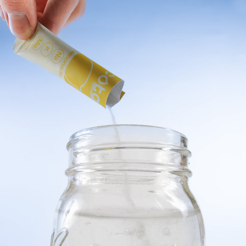 Hand pouring electrolyte powder from a lemonade flavored electrolyte stick packet into a clear glass jar against a light blue background.